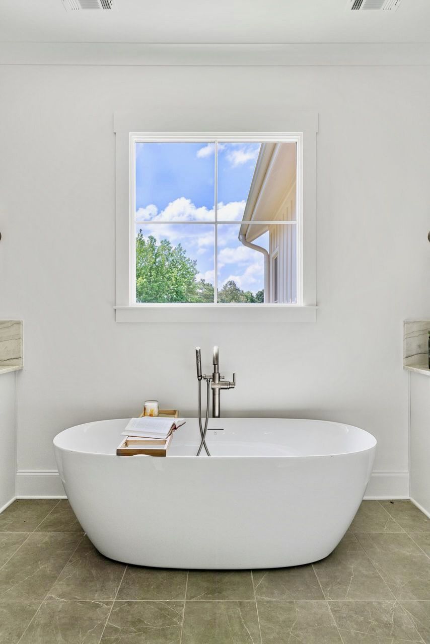 White bathtub in a modern bathroom with a window overlooking a blue sky and trees.