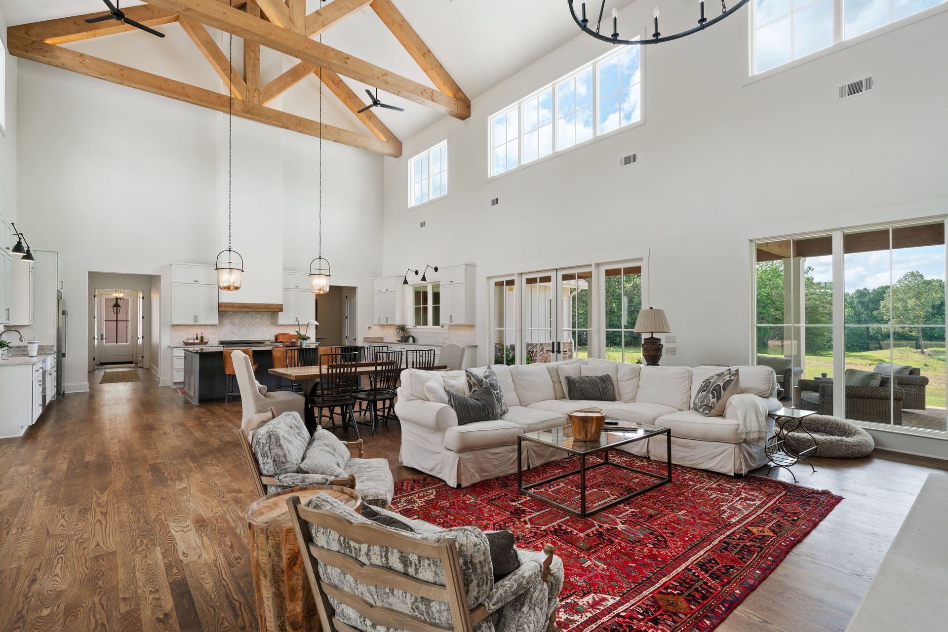 Spacious living room with wood beams, white walls, and red rug. Large windows overlook a green landscape.