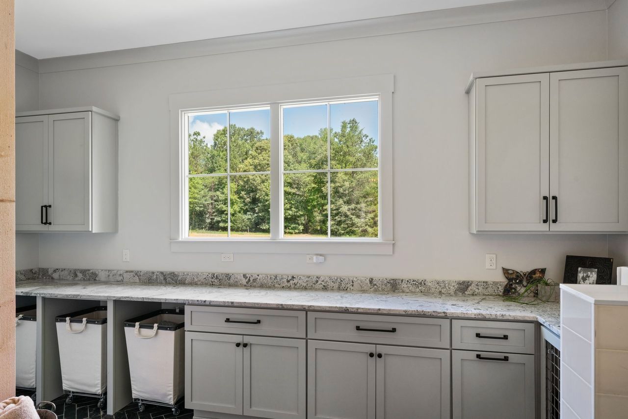Laundry room with grey cabinets, granite countertop, window with trees, and white laundry hampers.