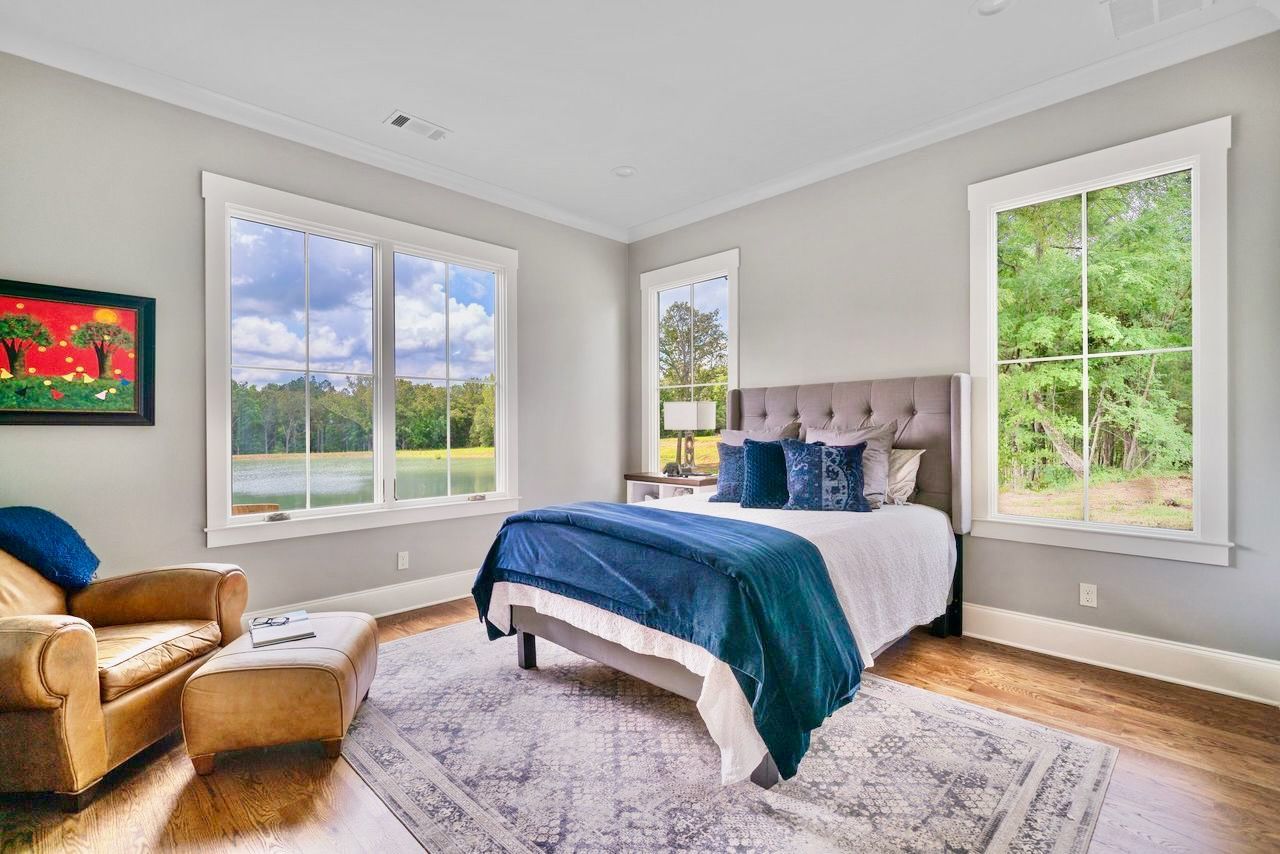 Bedroom with bed, armchair, large windows with lake view, painting on wall.