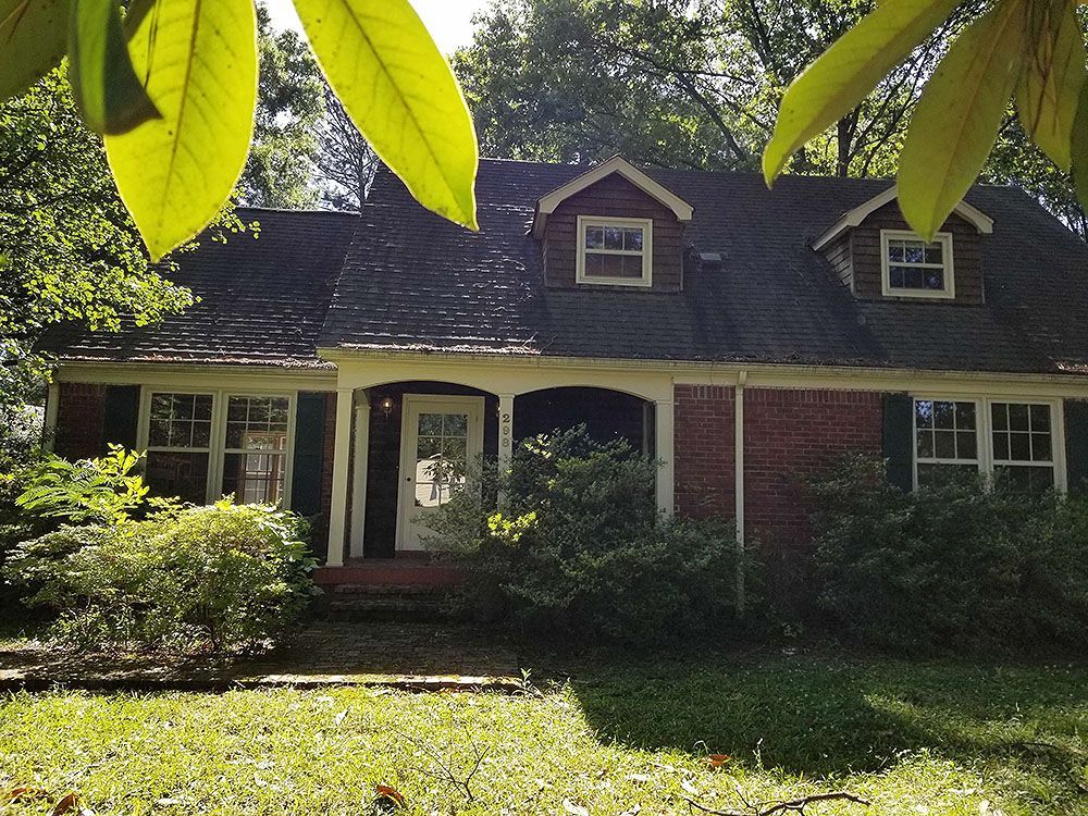 Exterior (Before) - Brick cottage house with green shutters, two dormers, and a porch framed by leafy trees.