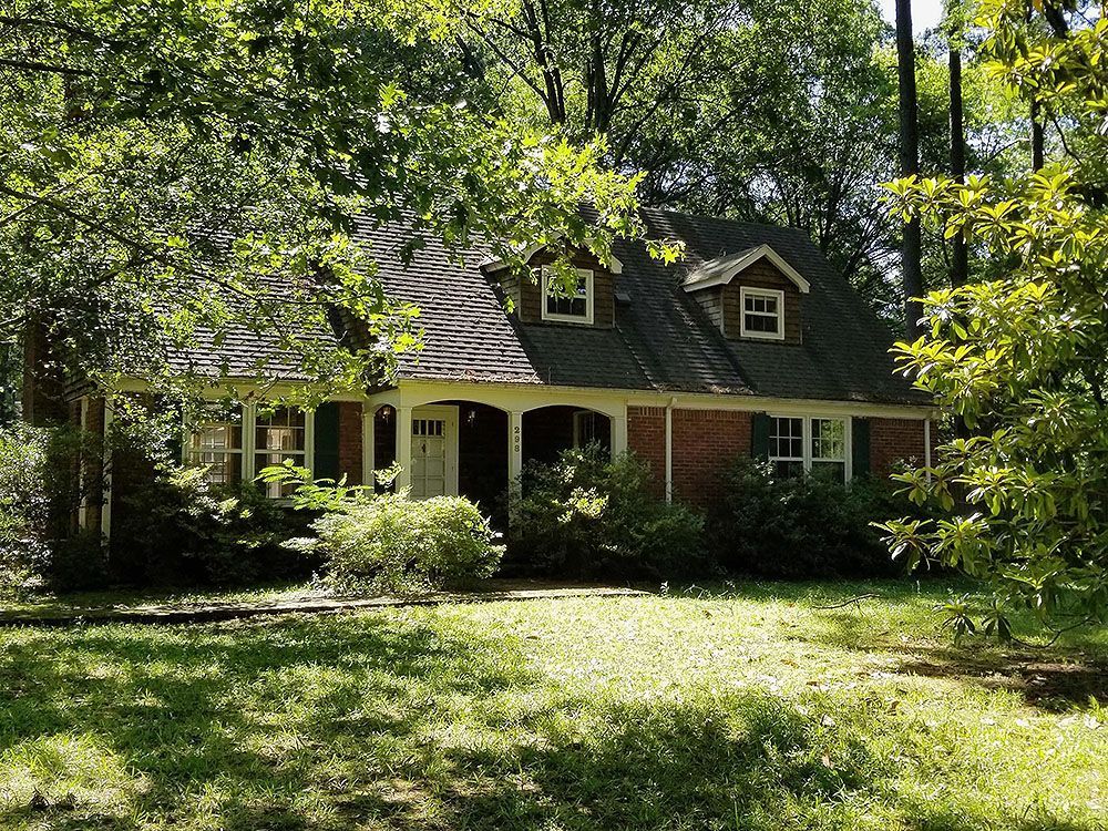 Exterior (Before) - Brick house with porch, dormers, and surrounding trees.