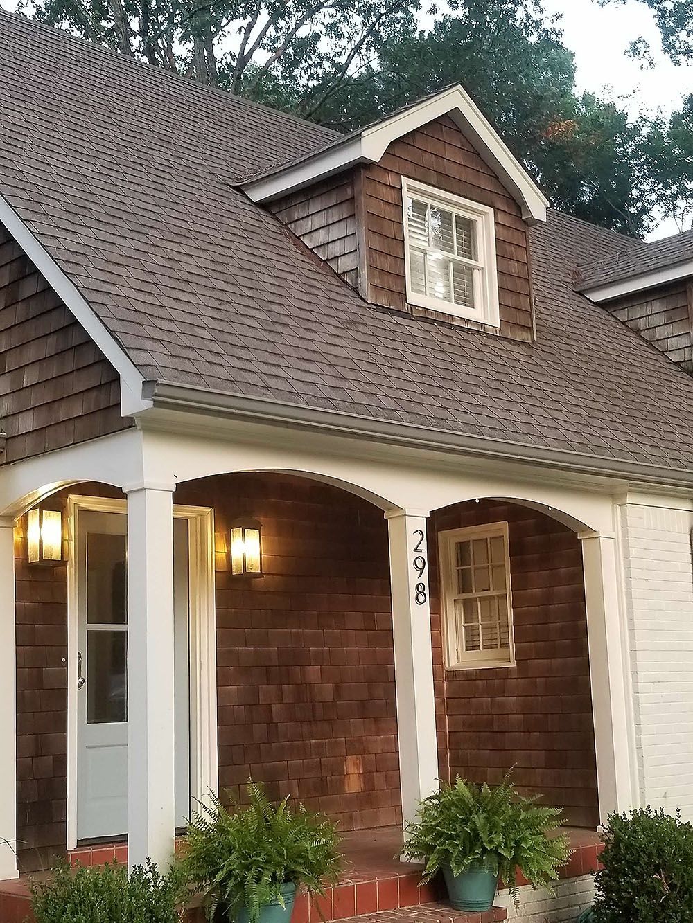 Front Door (After) - Brown shingle-covered house with white trim, arched porch, and dormer windows; potted plants in front.