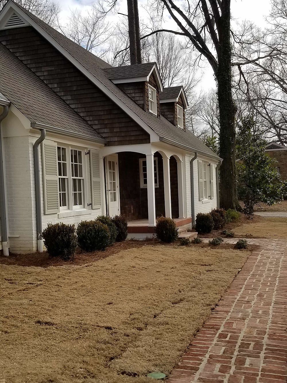 Front Door (In Progress) - White brick house with brown roof, shutters, and landscaping; red brick pathway.