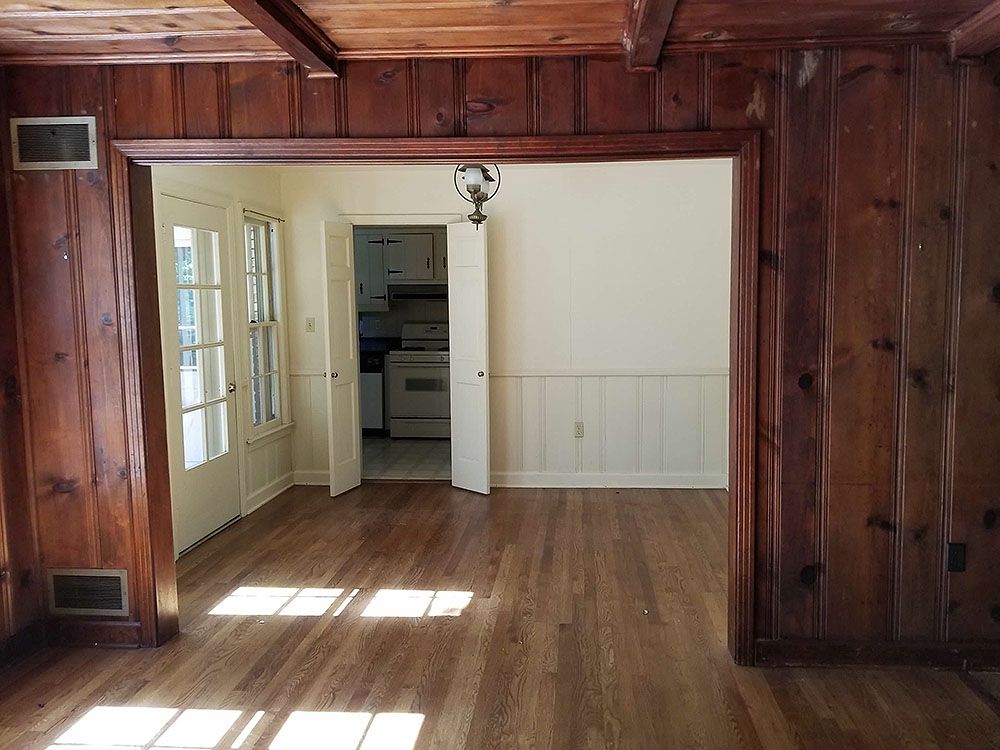 Kitchen (Before) - Wooden-paneled room with doorway, leading to kitchen. Hardwood floors and white wainscoting. Sunlit, empty interior.