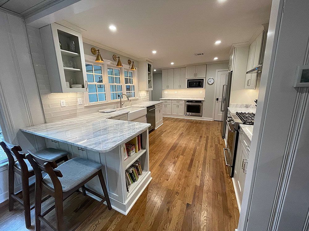 Kitchen (After) - A modern kitchen with white cabinets, marble countertops, and wooden floors. Bar stools are at the island.