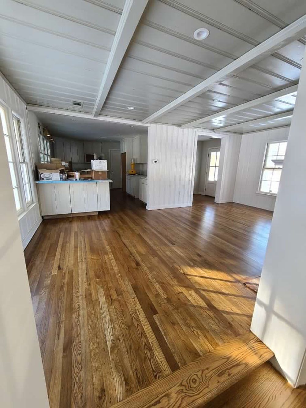 Kitchen (In Progress) - Newly renovated interior with hardwood floors, white ceiling beams, kitchen cabinets, and sunlight.