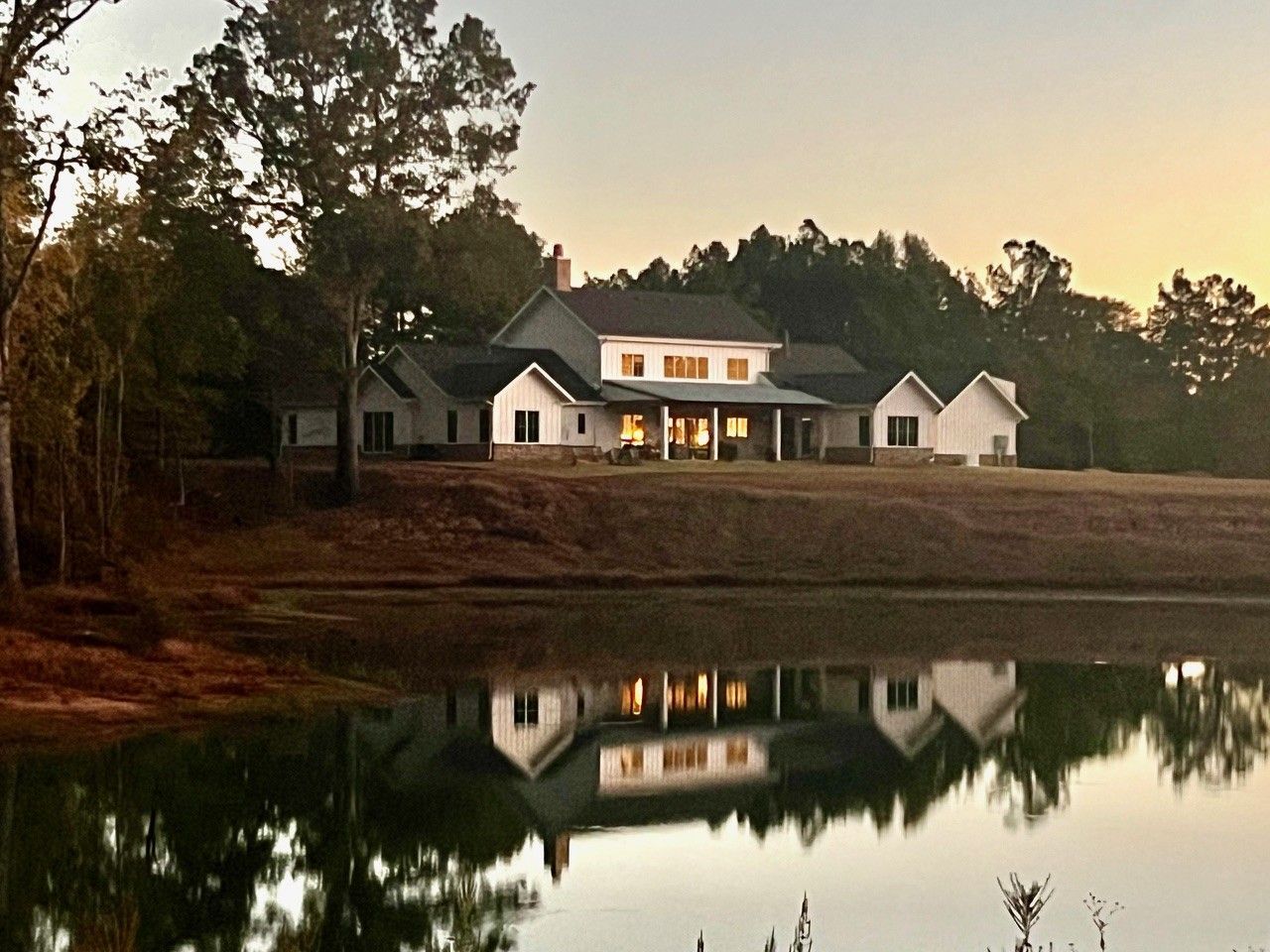 Large white lakeside house at dusk, reflected in calm water, surrounded by trees.
