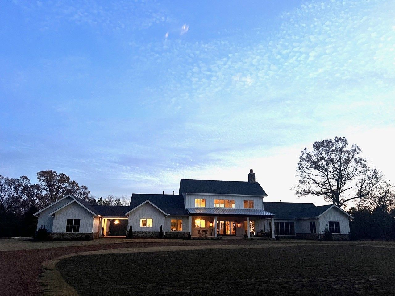Large white farmhouse at dusk with warm lights glowing under a blue evening sky