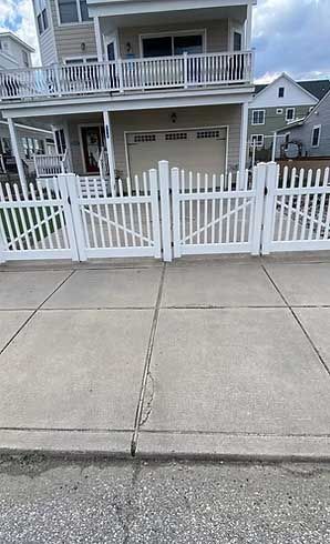 White picket fence with gate in front of a house, set on concrete sidewalk.