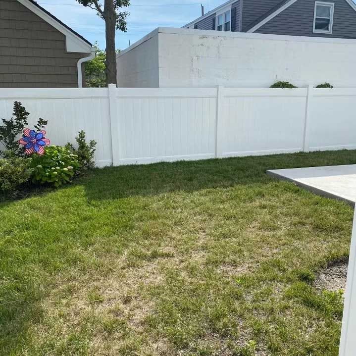 Backyard with white fence, green grass, and small flower garden under blue sky.