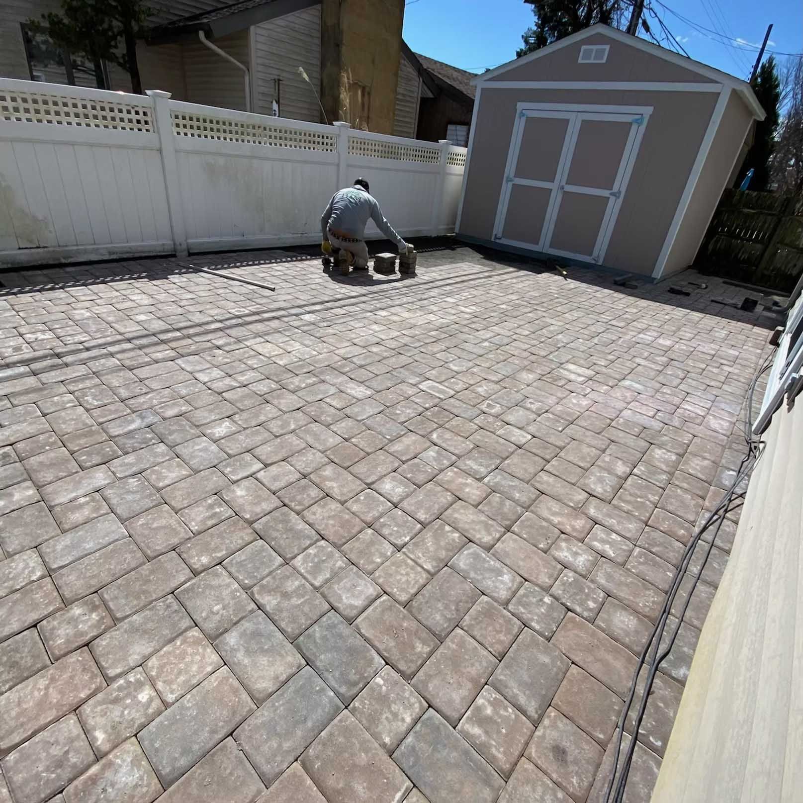 Person working on a new brick patio near a shed and white fence.