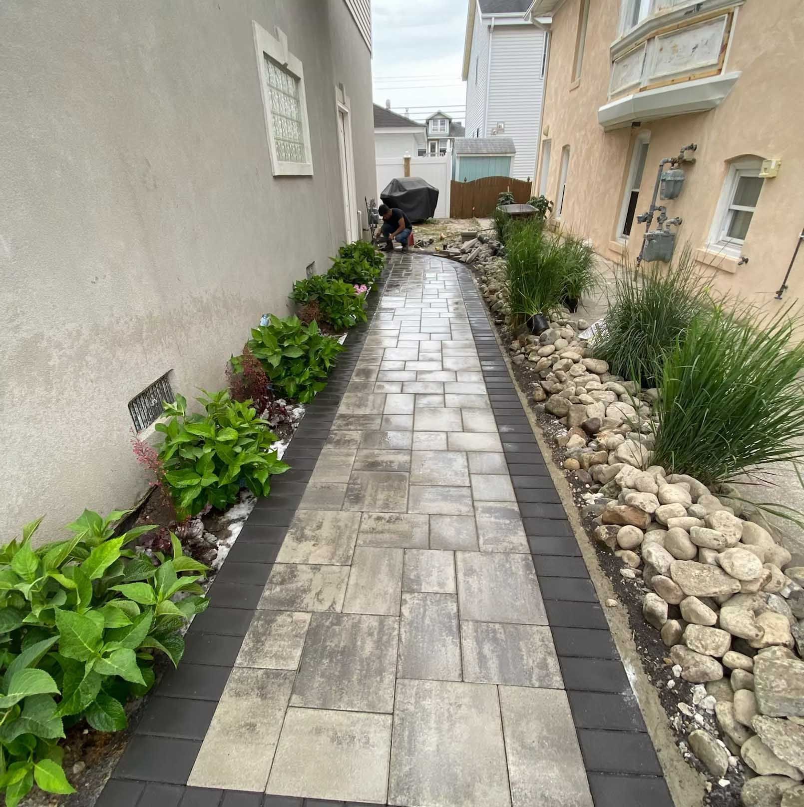 Narrow paved walkway between buildings, with plants and rocks along the sides.