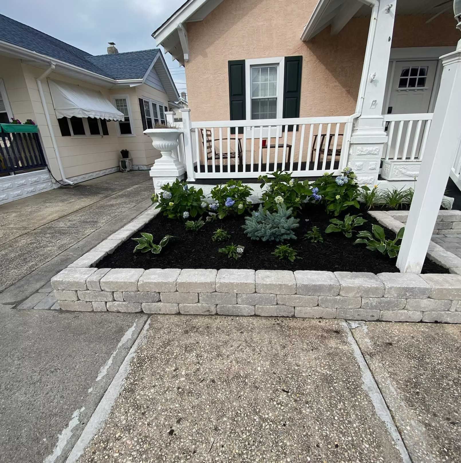 A front yard garden bed with a stone retaining wall and plants, in front of a house.