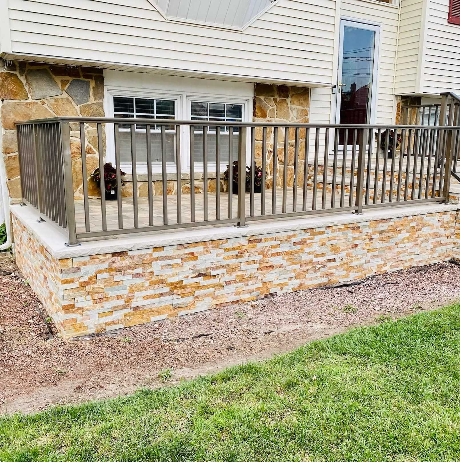 Stone-faced porch with metal railing and steps leading to a house entrance. Gravel and grass in the foreground.