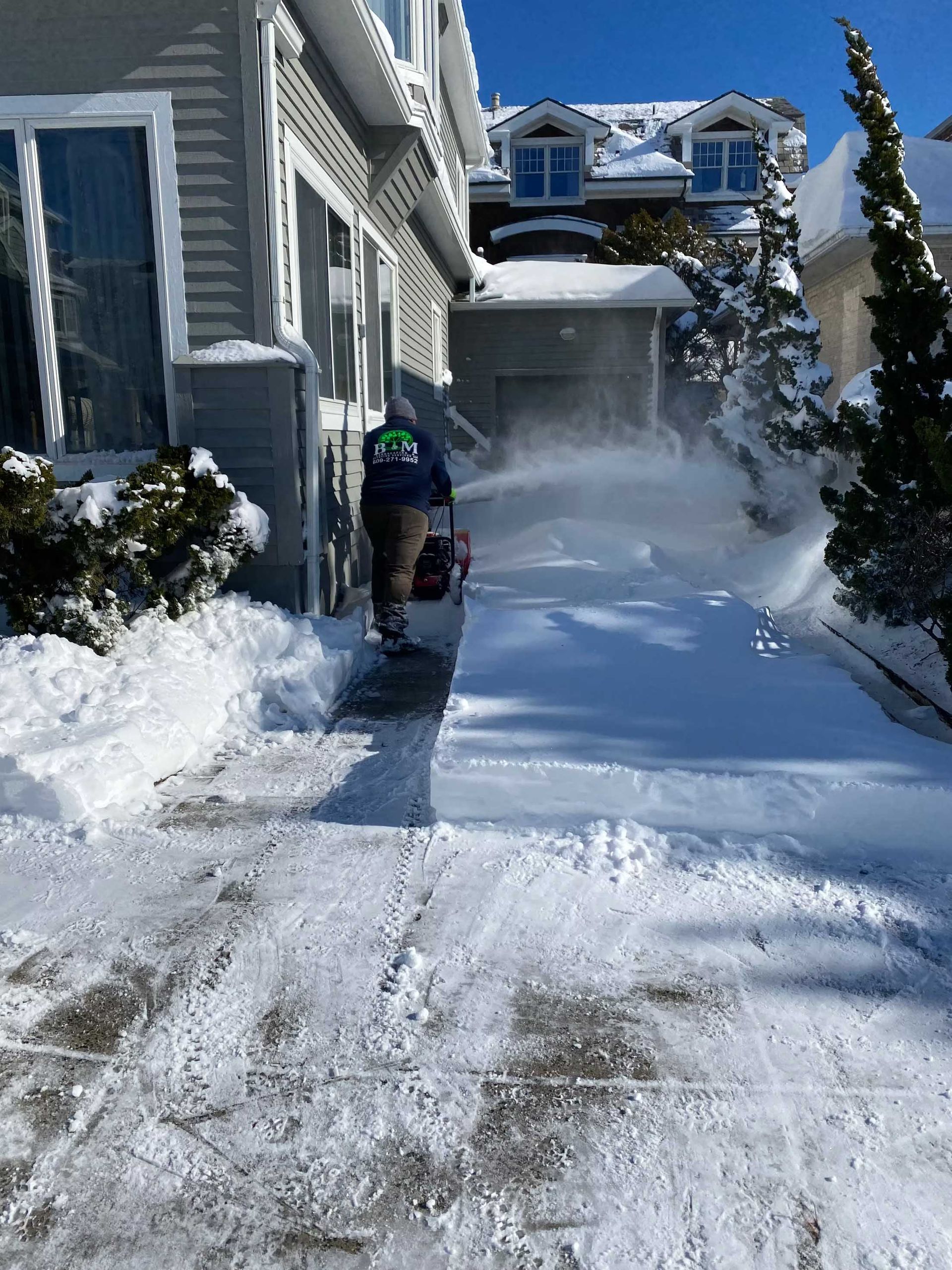 Person using a snow blower to clear snow from a driveway in front of a house on a sunny day.