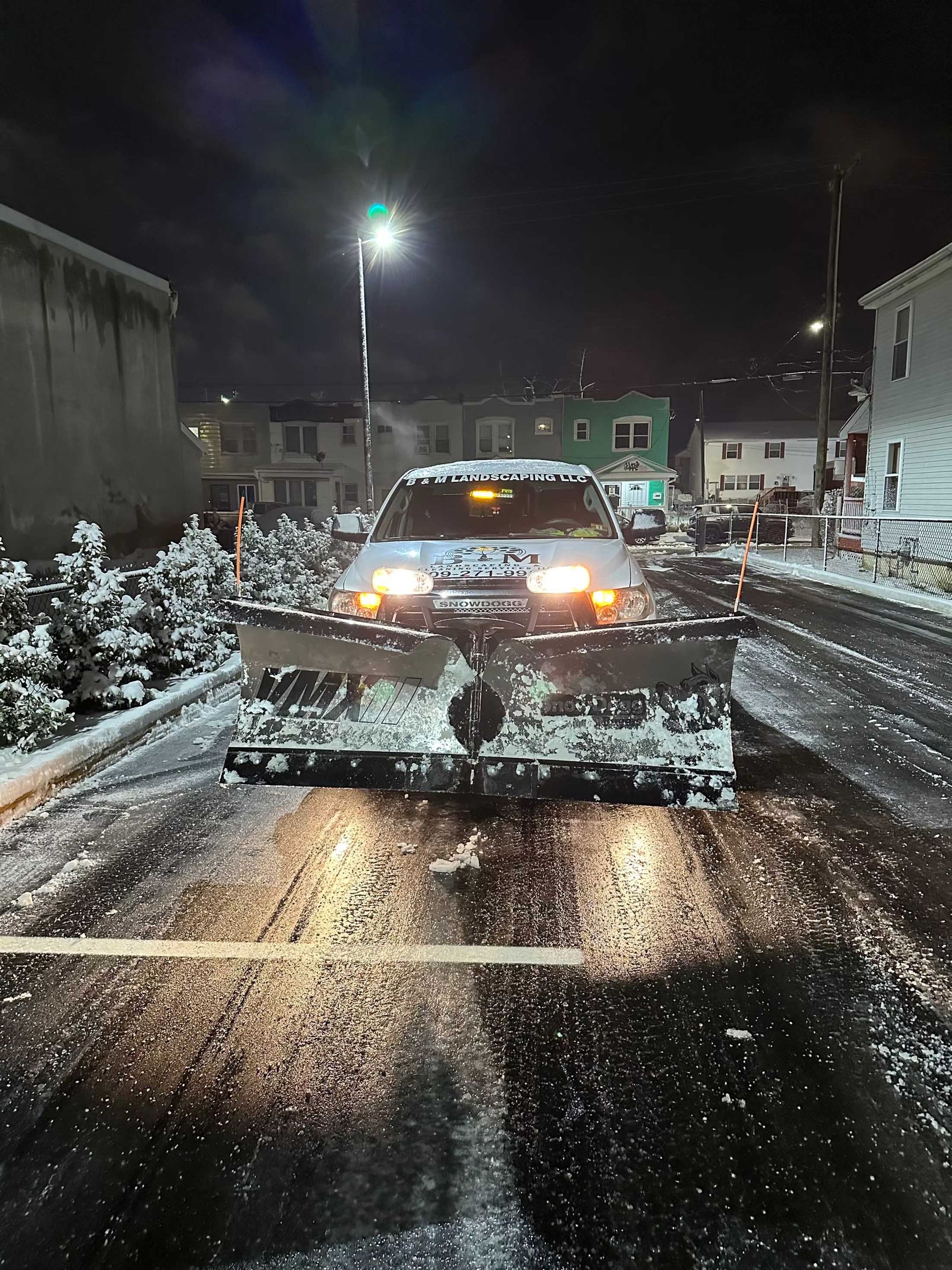 Snowplow clearing a snow-covered street at night with streetlights and buildings in the background.
