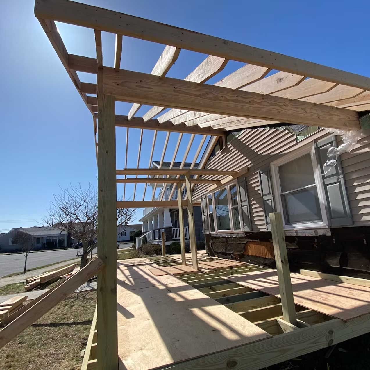 Wooden pergola under construction on a porch, with lumber and house in the background. Sunny day.