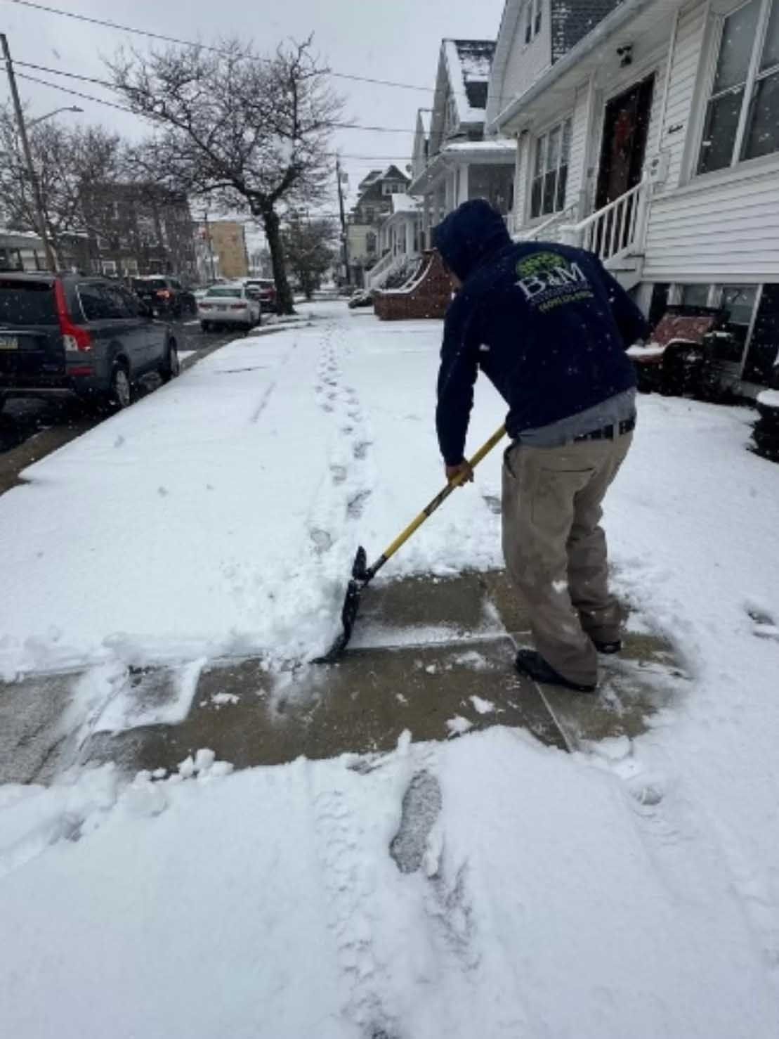 Person shoveling snow from a sidewalk in front of houses on a snowy day.
