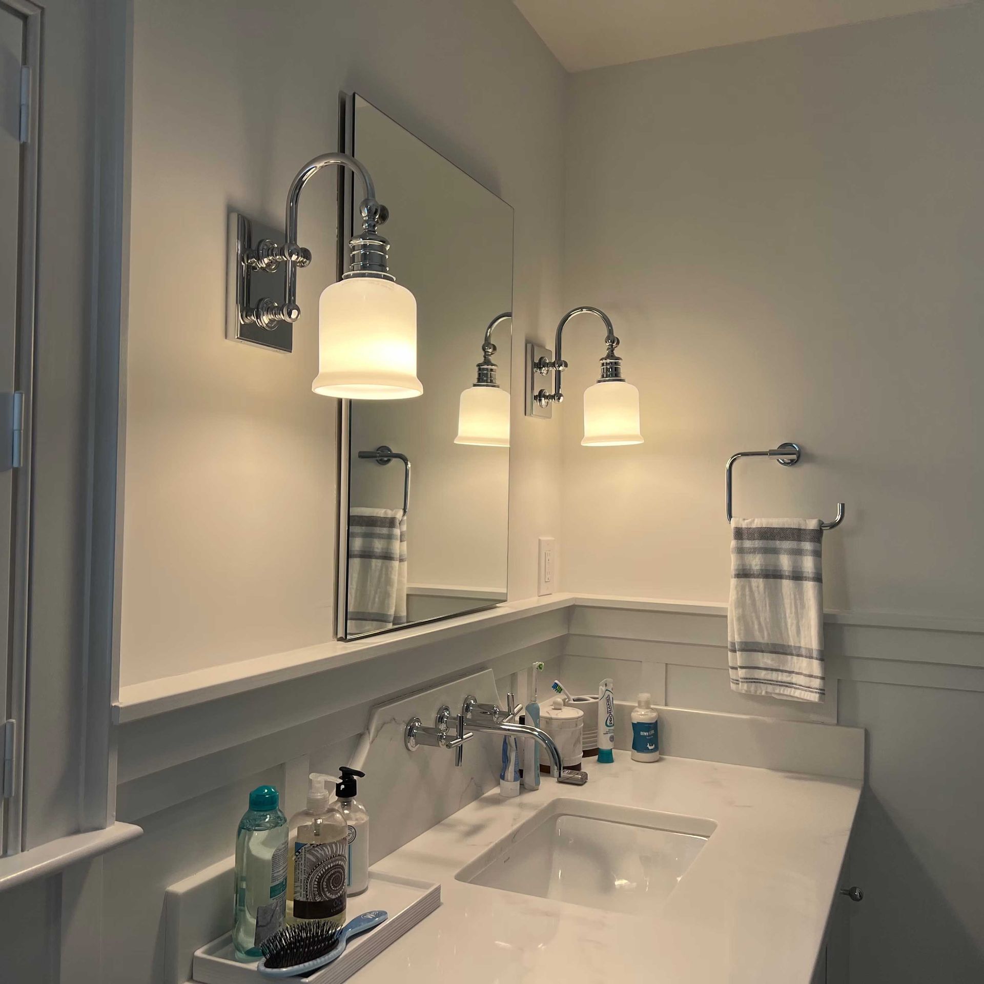 Bathroom with a white countertop, sink, and mirror.  Wall-mounted lights flank the mirror. A towel rack with a towel is visible.