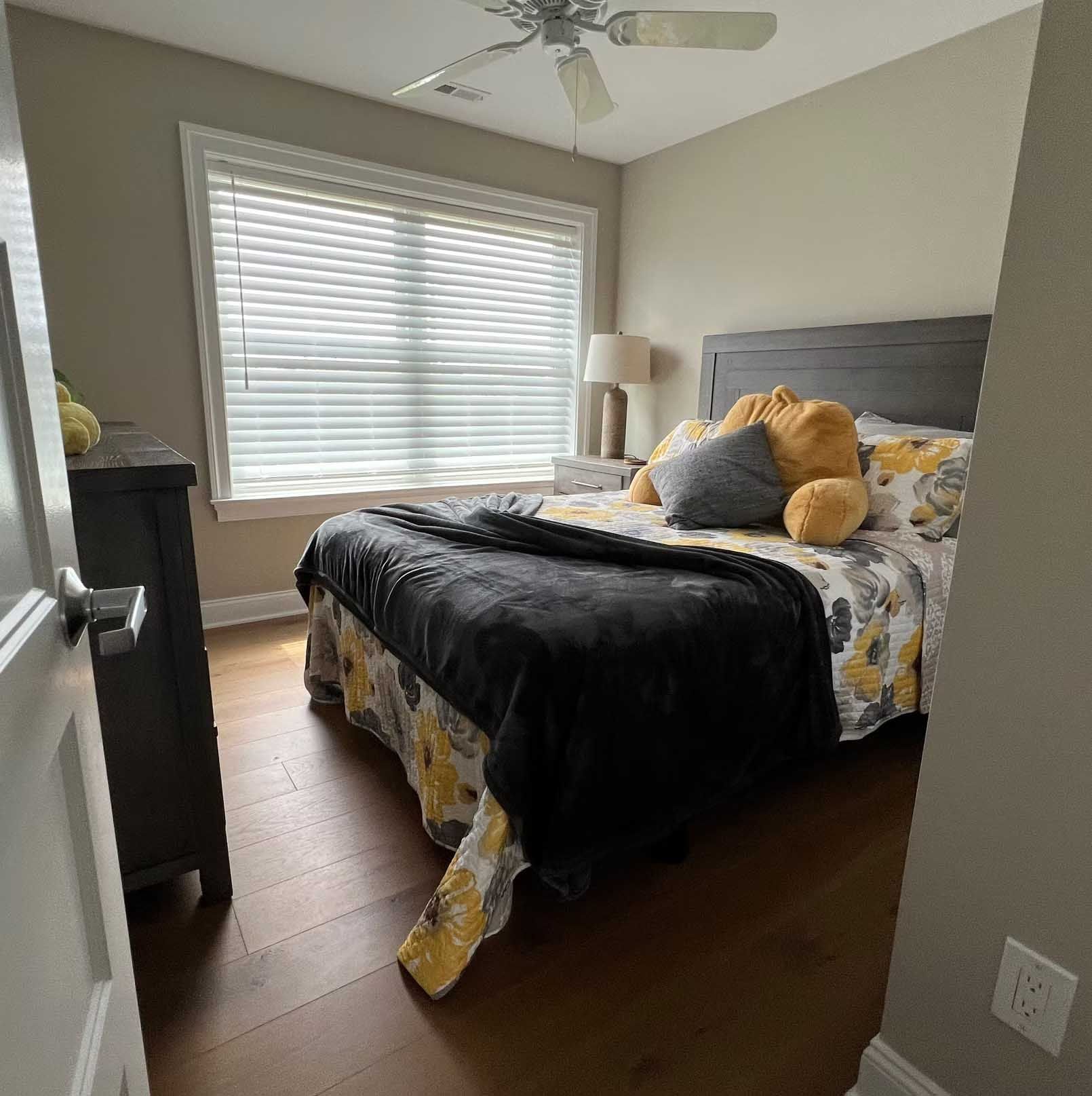 Bedroom with bed, dresser, window with blinds, and ceiling fan. The bed has yellow and black bedding.
