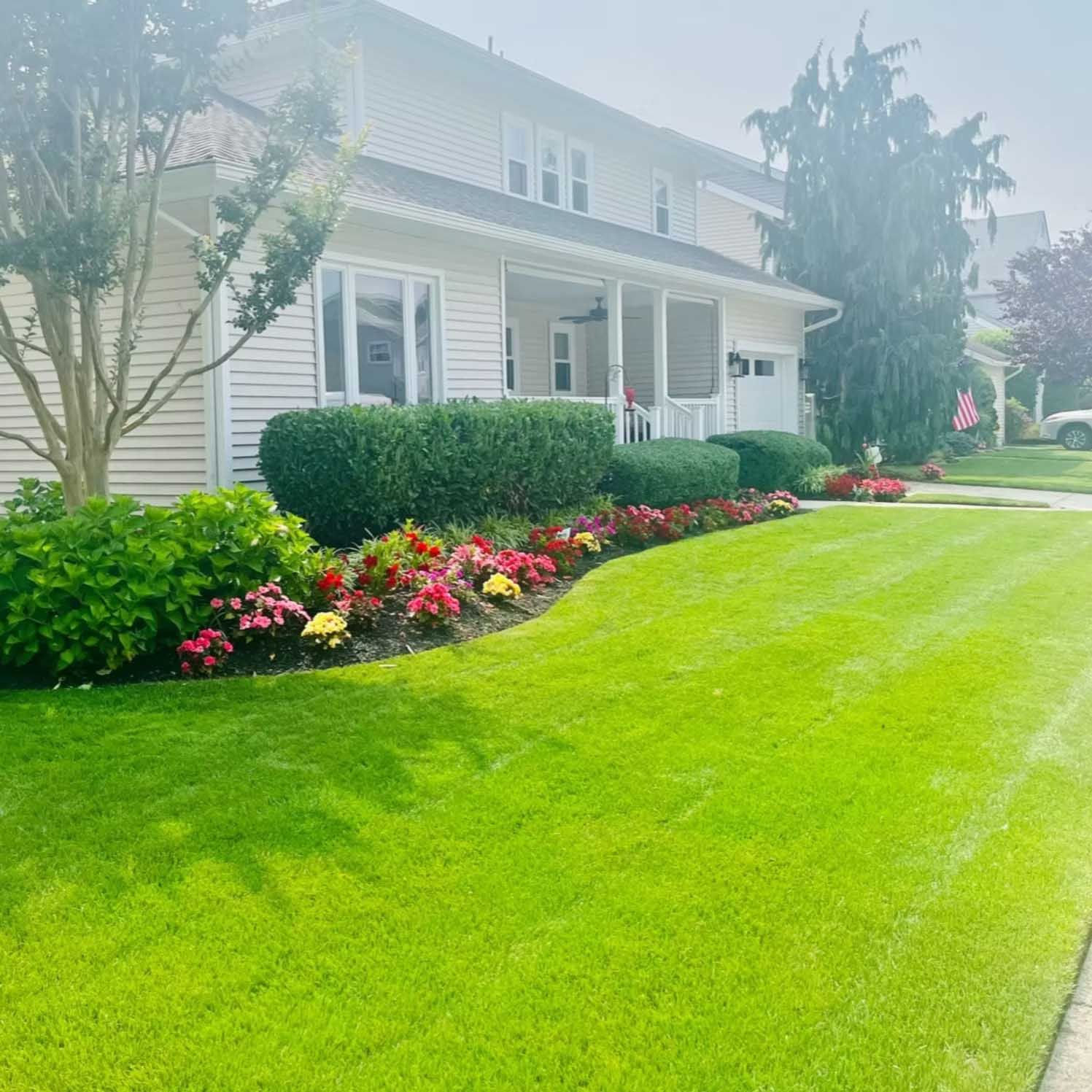 Well-manicured lawn with flowerbeds in front of a two-story beige house with a porch and a garage.