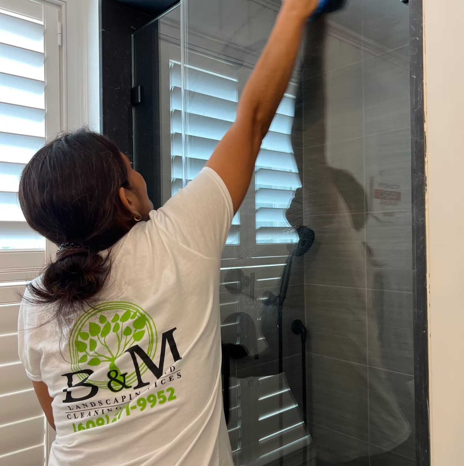 Woman cleaning a glass shower door. She wears a white shirt with a logo and has her hair in a ponytail.