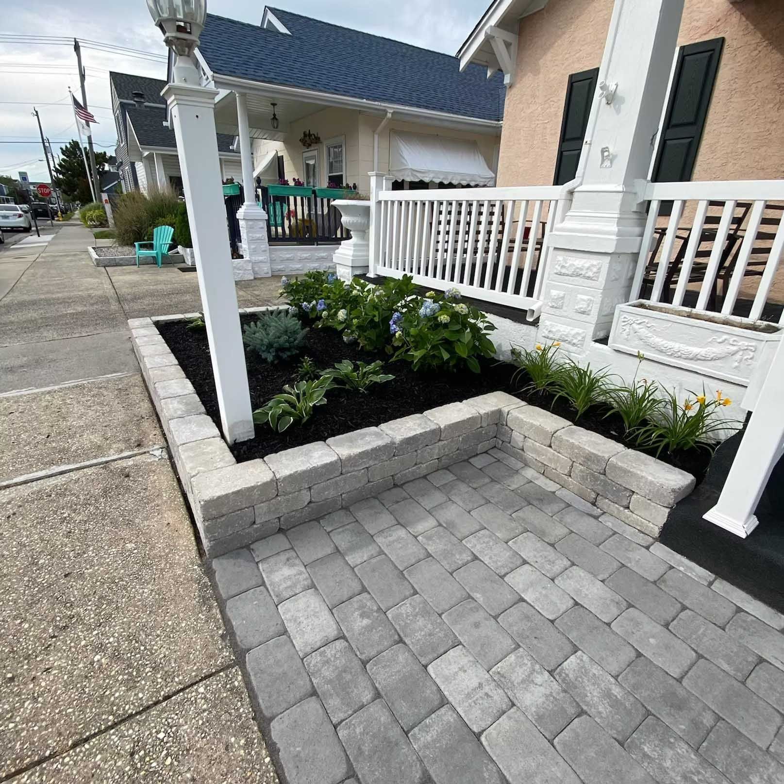 Stone-paved walkway and raised garden bed in front of a house. White porch, blue hydrangeas, dark mulch.