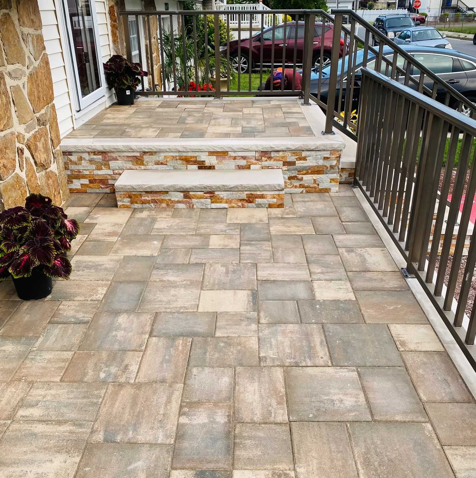 Stone paved patio with steps leading to a raised porch, flanked by metal railings, with potted plants.