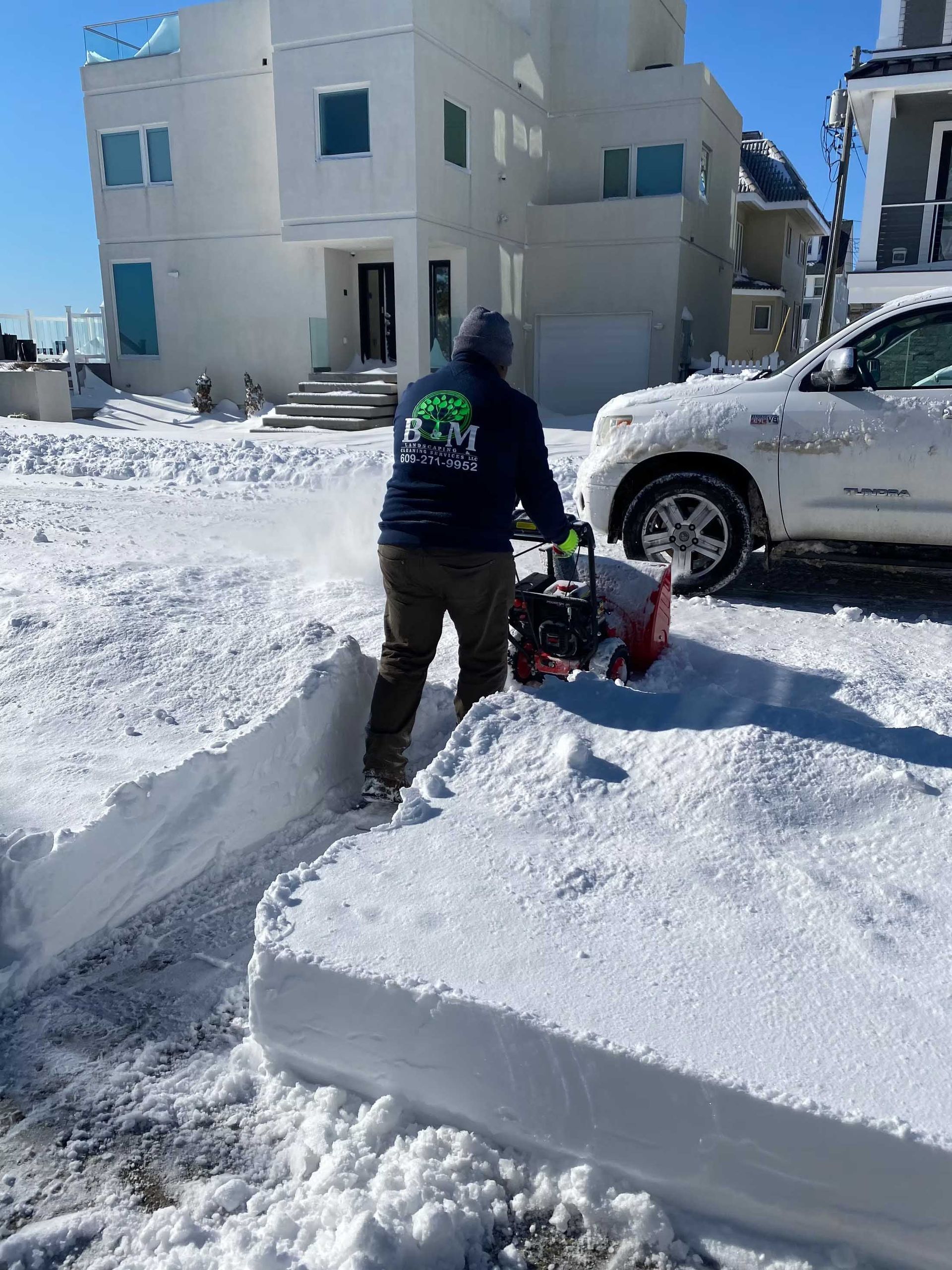 Person using a snow blower to clear a snowy driveway in front of a white house.