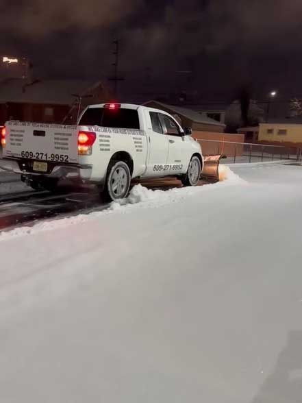 White pickup truck plowing snow at night. The truck has a snowplow blade and business information on the sides.