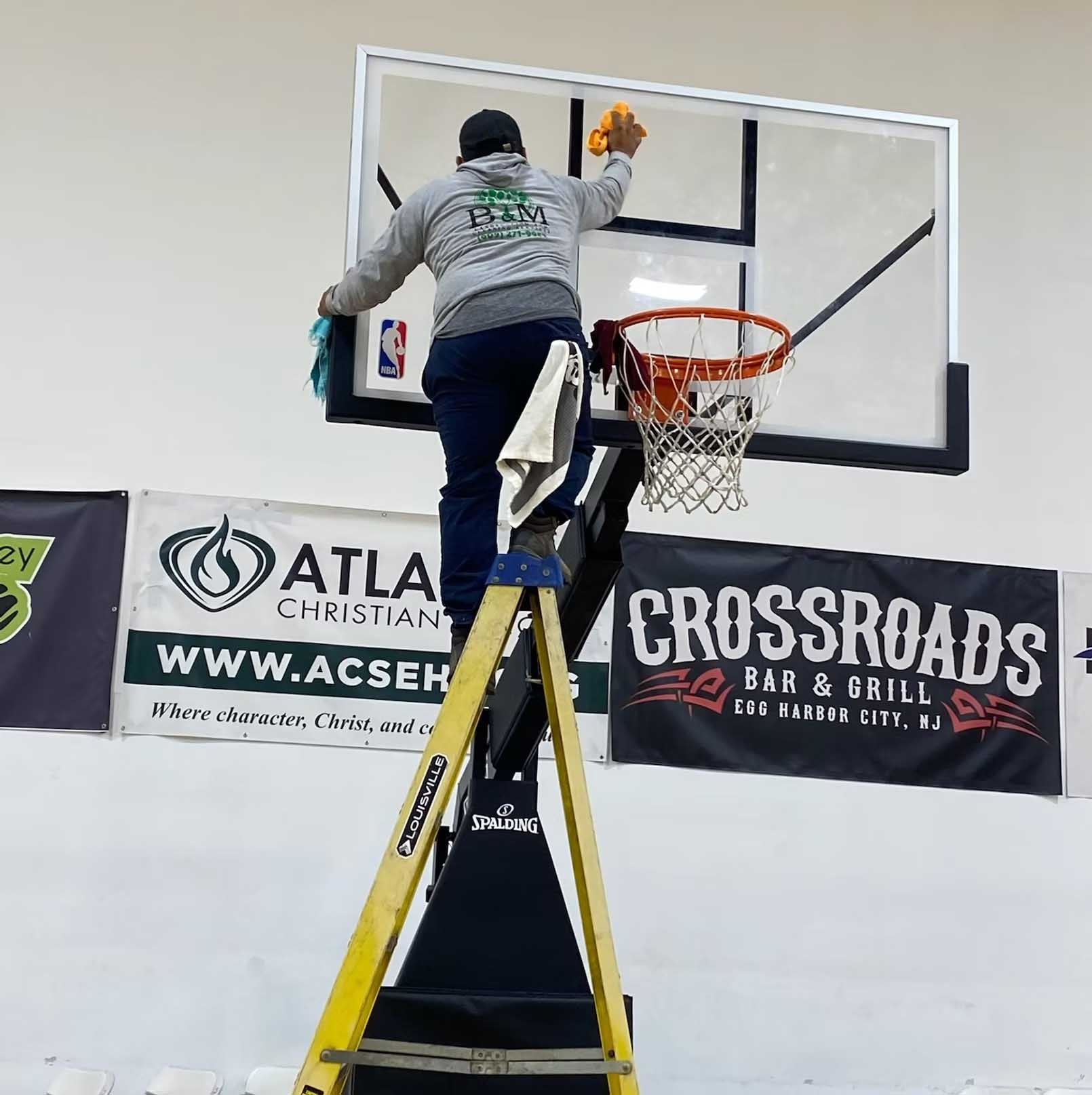 Man on a ladder cleaning a basketball hoop in a gym.