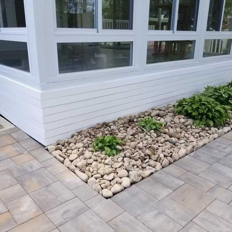 White-sided building with window, bordered by a stone garden bed and paved patio.
