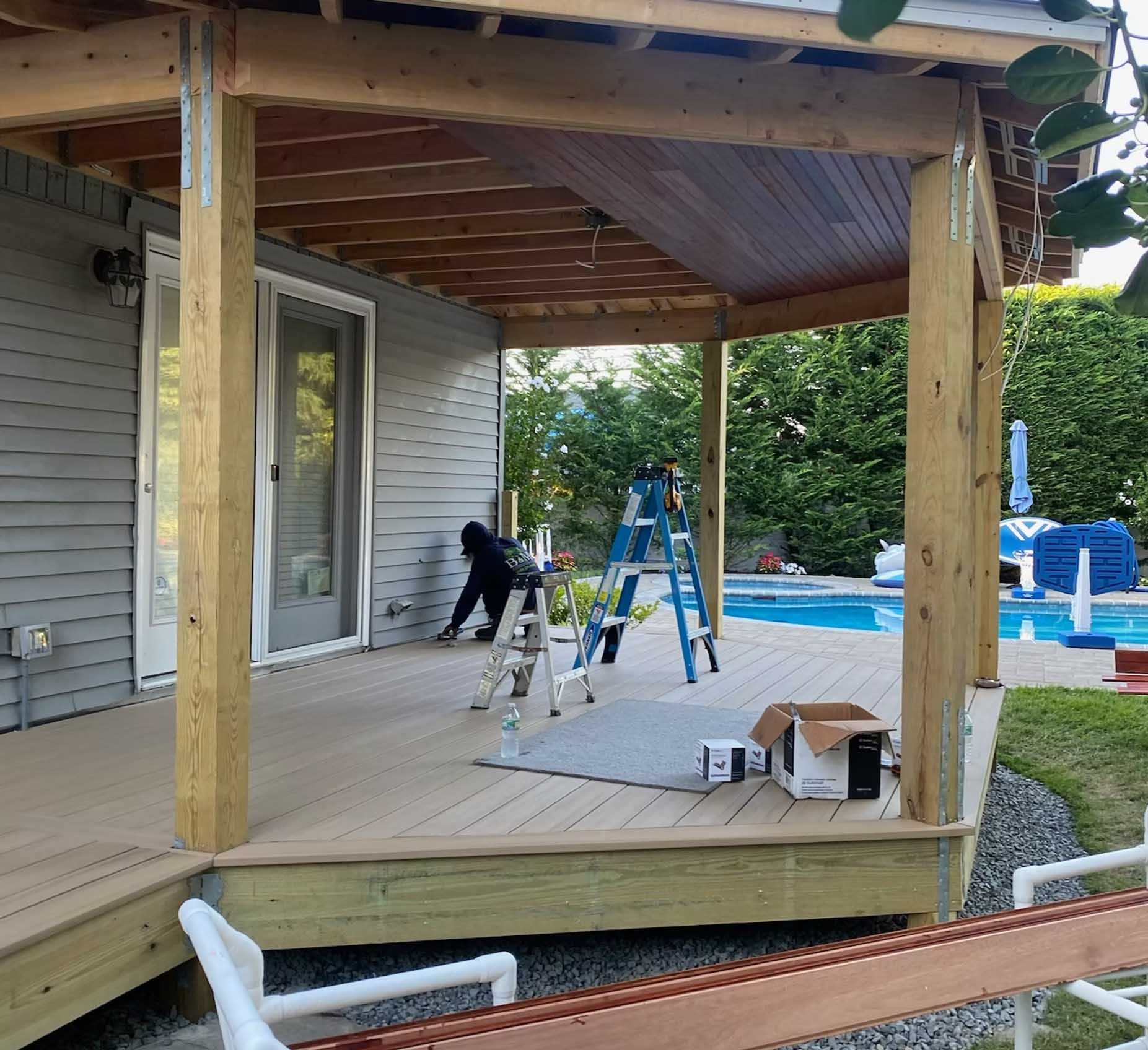 A person works on a partially built deck with a covered area. Pool in the background.