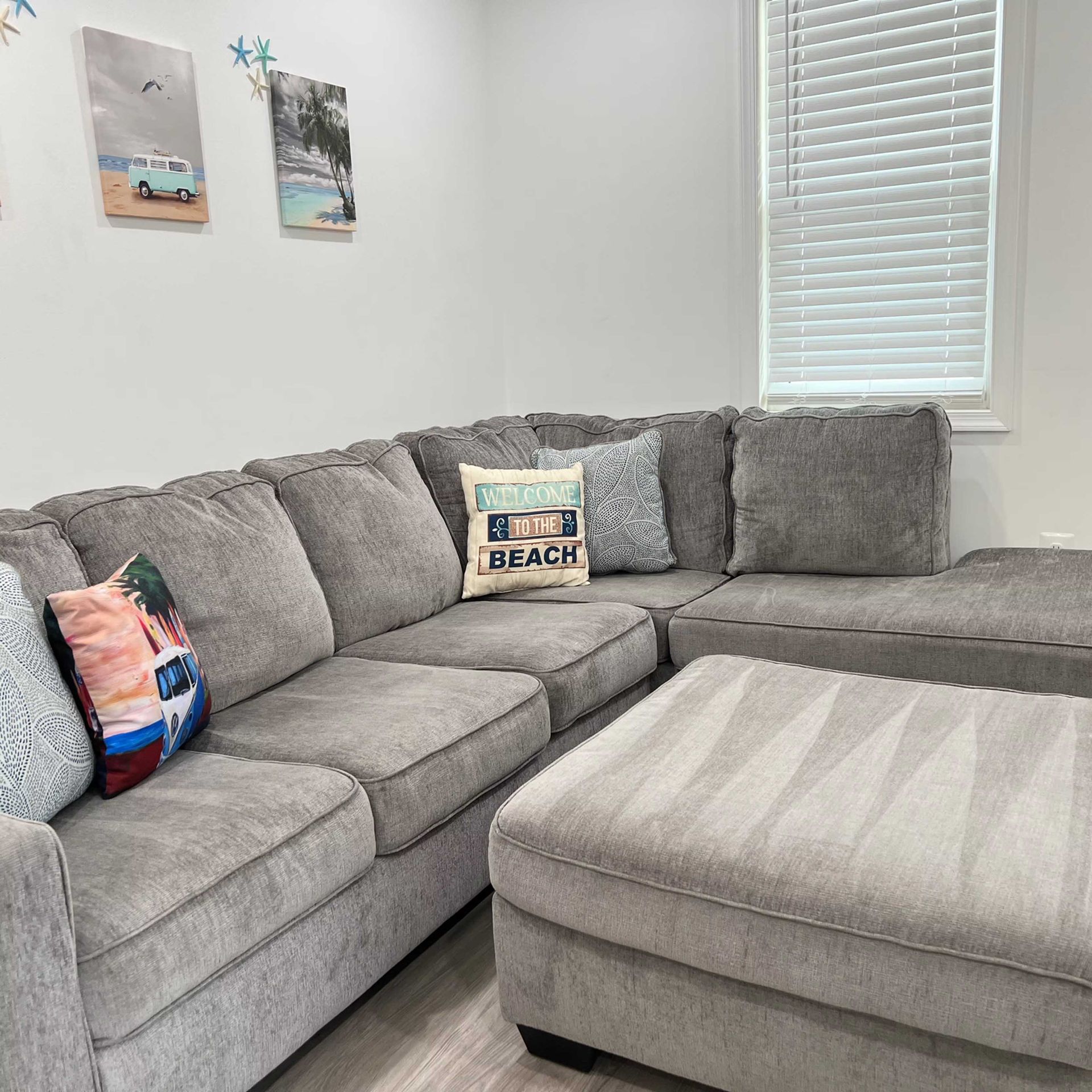 Gray sectional sofa with ottoman and beach-themed pillows. White walls, framed artwork, and window.