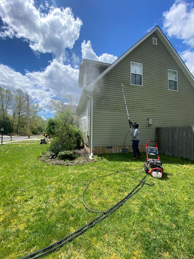 Person pressure washing the green siding of a house with a long wand. Red pressure washer on the lawn.