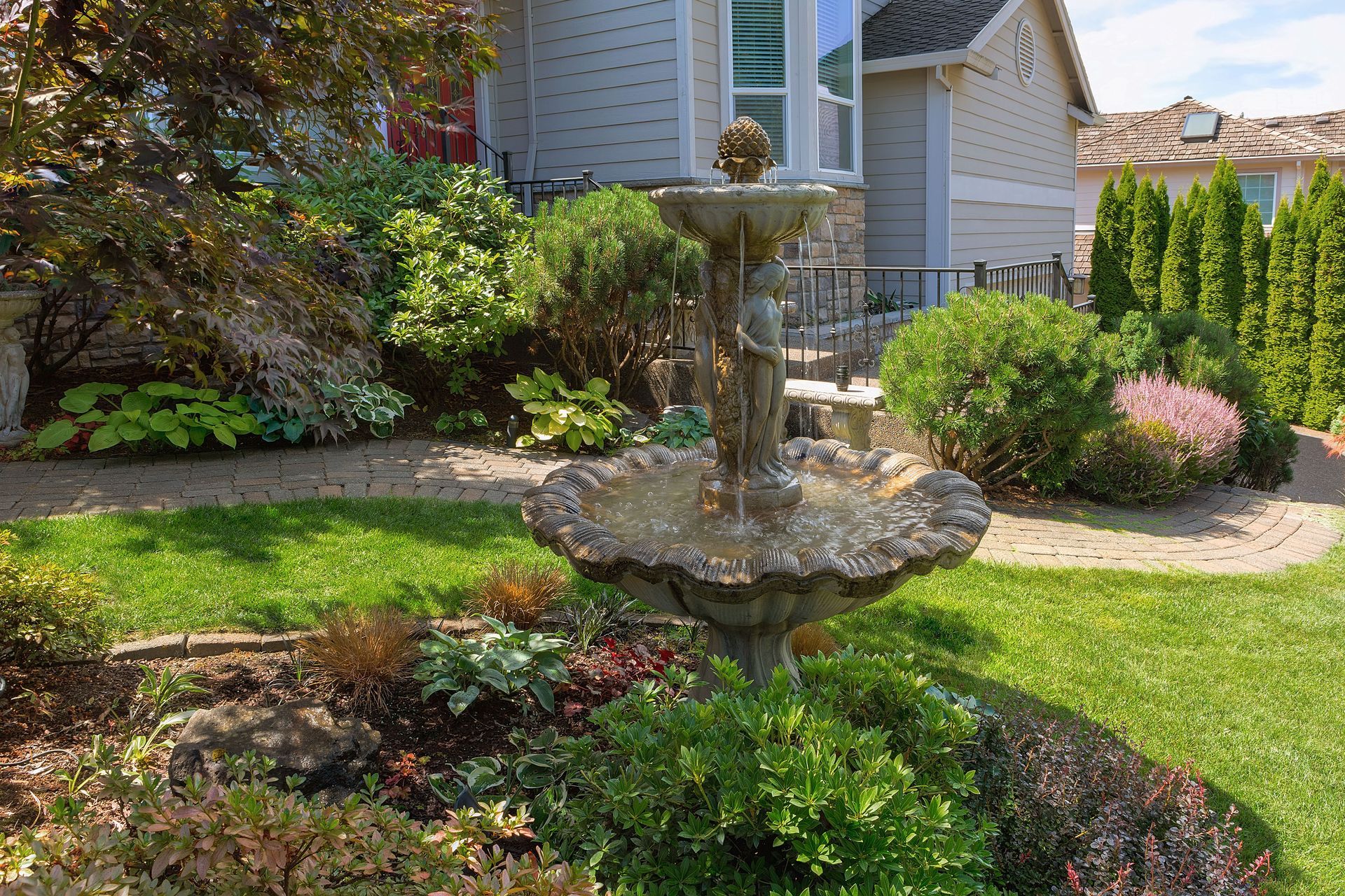 Ornamental fountain in a garden bed with manicured green grass in front of a house.