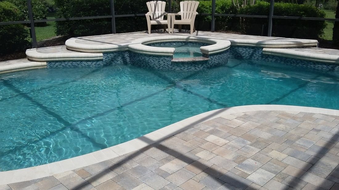 A swimming pool with an attached hot tub, surrounded by stone pavers, enclosed in a screened-in lanai.