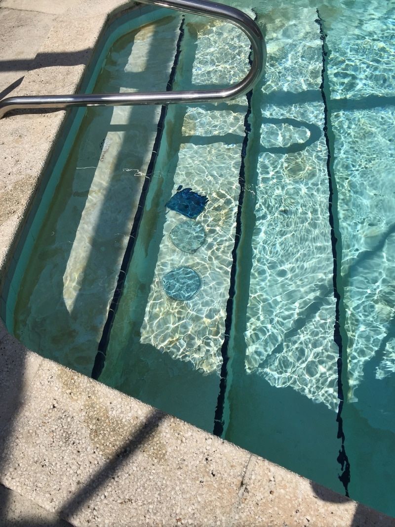 Concrete pool steps leading into turquoise water, with a metal handrail and small dark circular drain covers visible.
