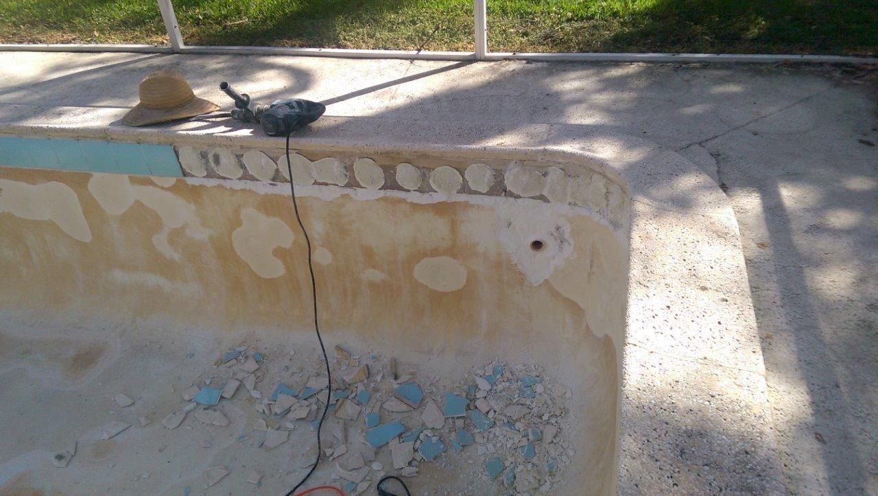 A dry, drained swimming pool with crumbling decorative tiles along the upper edge and construction tools on the deck.