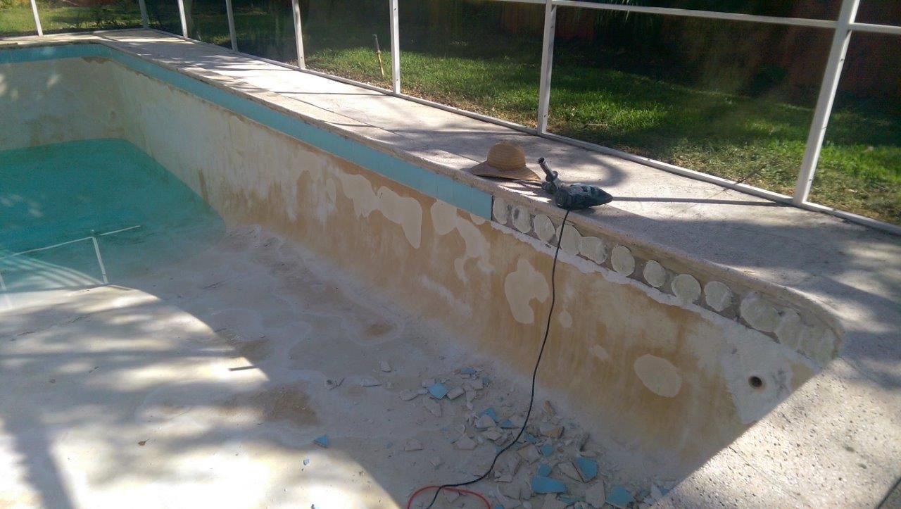 A pool deck with a partially removed blue tile waterline, power tools, and debris visible on the concrete edge.