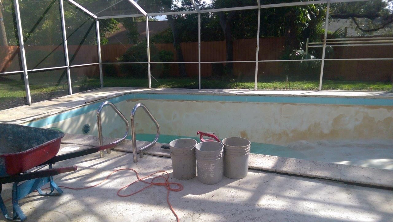 An empty backyard swimming pool undergoing renovation, with a wheelbarrow and three buckets sitting on the patio deck.