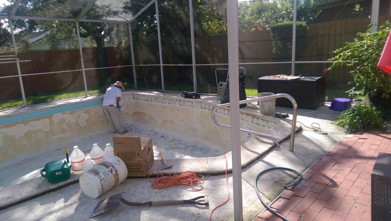 A worker performs maintenance on an empty backyard swimming pool under a screened enclosure.