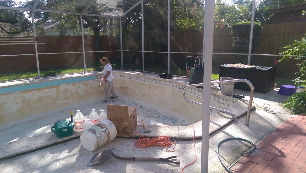 A person stands inside an empty, light-colored swimming pool surrounded by a screen enclosure, with supplies and tools.