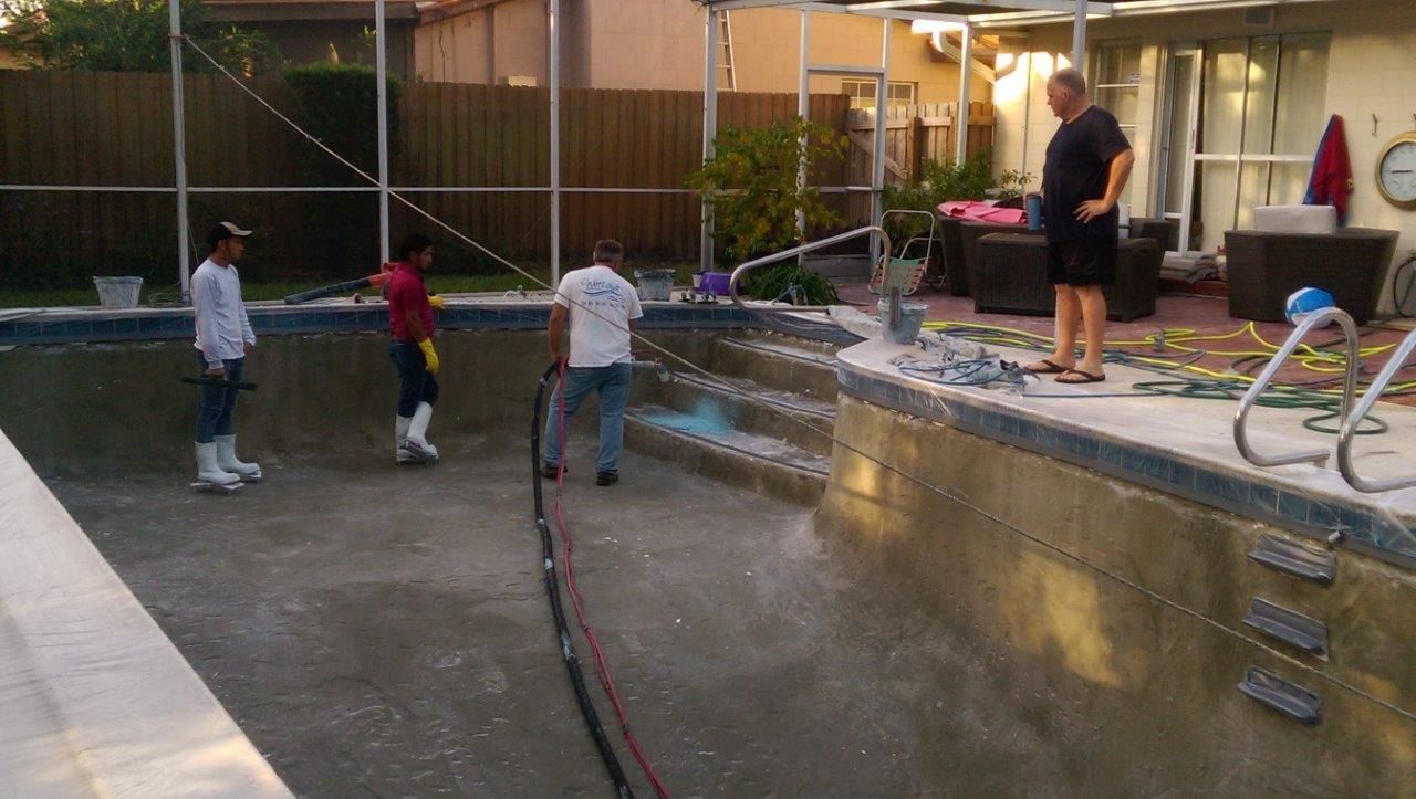Three people work on cleaning and repairing an empty backyard swimming pool while a fourth person watches.