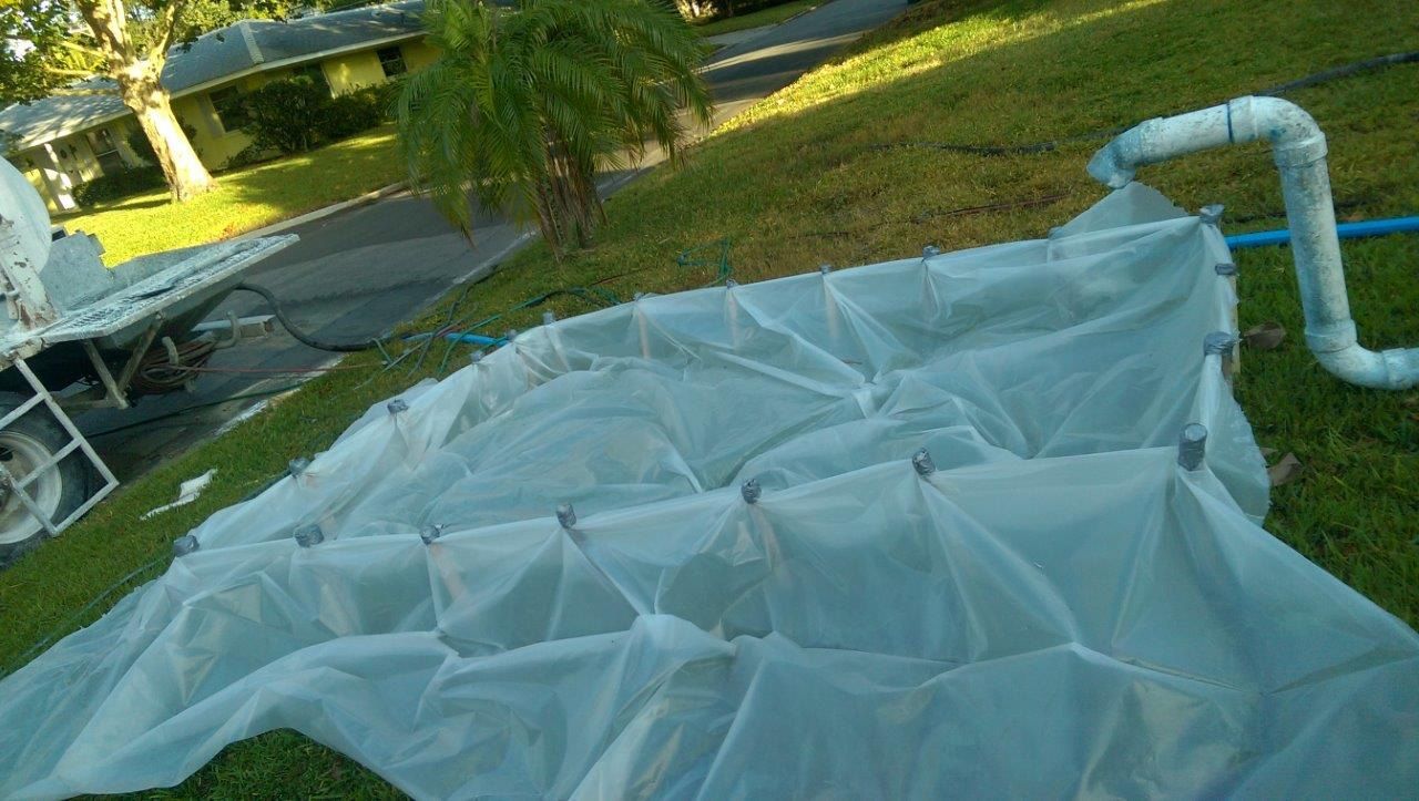 A white mesh fabric is draped over a rectangular frame on a grassy lawn near a white plastic pipe structure.