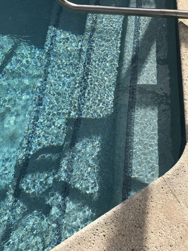 Top-down view of turquoise swimming pool water with submerged concrete steps and a metal railing.