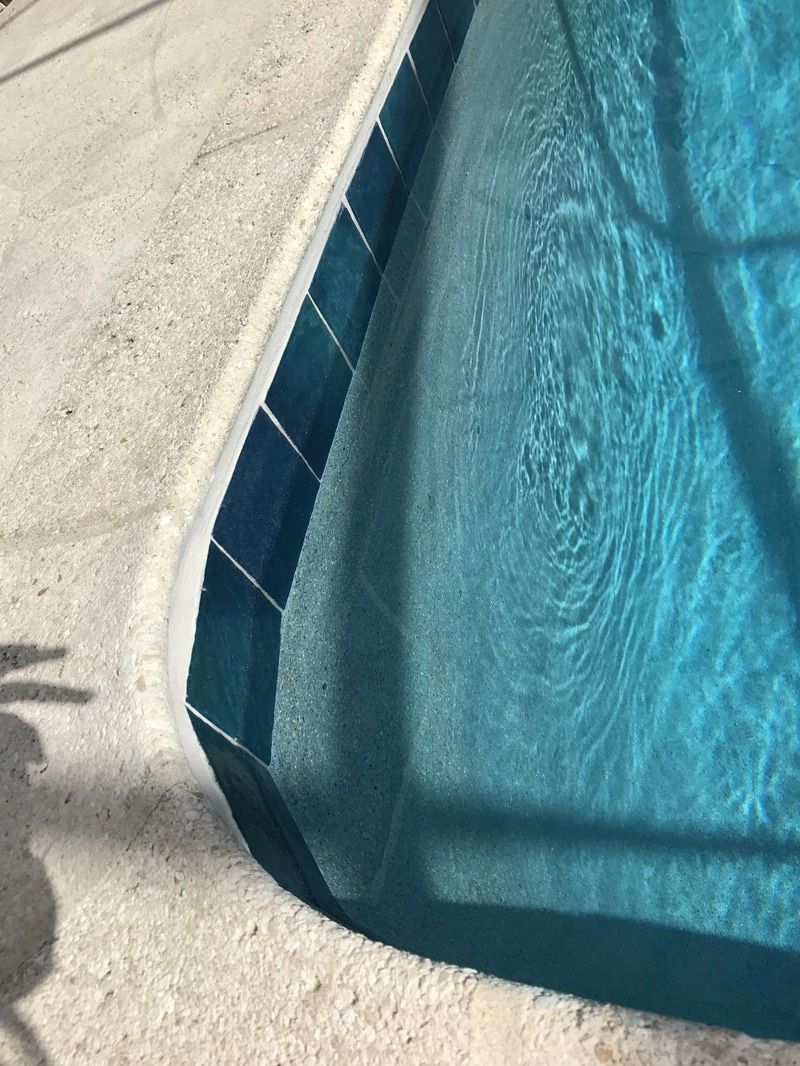 Close-up view of a swimming pool corner featuring blue border tiles and light-colored textured pool deck concrete.