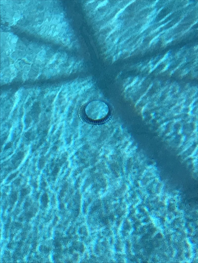 A small, circular pool drain sits on the blue tiled floor of a swimming pool, partially obscured by long, dark shadows.