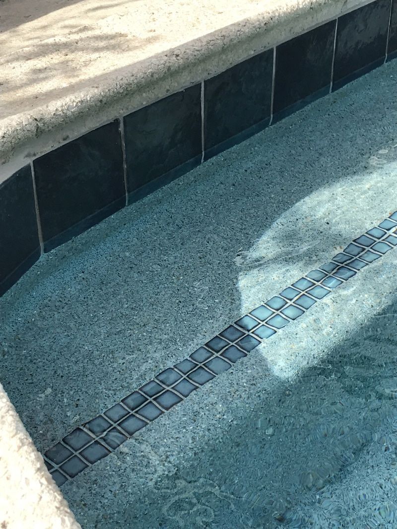 A swimming pool edge featuring dark square waterline tiles, a light concrete coping, and a step with decorative inlay.